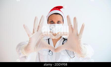 Ärztin mit einer medizinischen Maske und Händen in Latexhandschuh zeigt das Symbol des Herzens. Arzt für das Herz. Arzt mit Weihnachtsmann Hut. Stockfoto