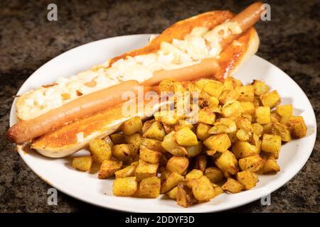 Ein spanischer Hot Dog serviert mit Patatas Bravas. Die Hot Dogs wurden in einem Baguette gebacken und mit Käse und gehackten Zwiebeln überbacken. Stockfoto