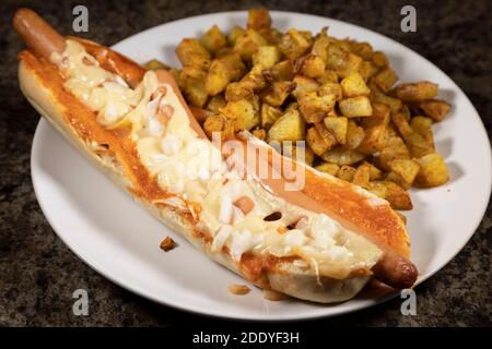 Ein spanischer Hot Dog serviert mit Patatas Bravas. Die Hot Dogs wurden in einem Baguette gebacken und mit Käse und gehackten Zwiebeln überbacken. Stockfoto