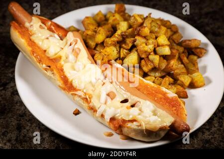 Ein spanischer Hot Dog serviert mit Patatas Bravas. Die Hot Dogs wurden in einem Baguette gebacken und mit Käse und gehackten Zwiebeln überbacken. Stockfoto