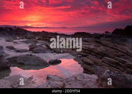 Ein leuchtender roter Sonnenuntergang über einem Strand, mit den Wolken, die sich in den Felsenpools spiegeln Stockfoto