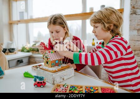 Kinder dekoriert Weihnachten Lebkuchenhaus mit bunten Zuckerguss und Süßigkeiten auf dem Tisch in der Küche. Weihnachten Familie Home Aktivitäten concep Stockfoto