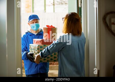 Lieferer bringt Urlaubspakete. Frau zu Hause stehend in der Tür, Empfang Pakete für Weihnachtsgeschenke. Delivery guy in Schutzmaske Stockfoto