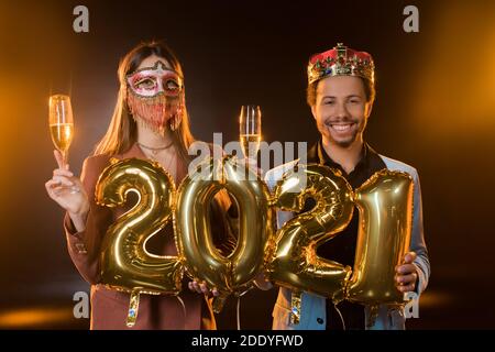 Glücklicher afroamerikanischer Mann in Krone hält Ballons mit 2021 Zahlen bei Frau in Karnevalsmaske mit Glas Champagner Auf Schwarz Stockfoto