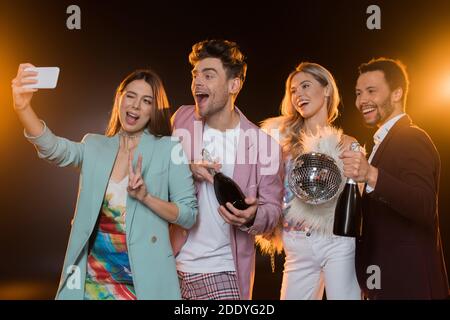 Glückliche multikulturelle Männer halten Flaschen mit Champagner in der Nähe von Frauen während Selfie auf Schwarz Stockfoto