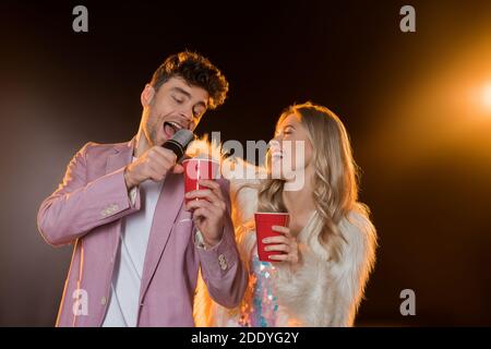 Mann singen, während das Mikrofon in der Nähe der Frau mit Kunststoff-Tasse halten Auf Schwarz Stockfoto