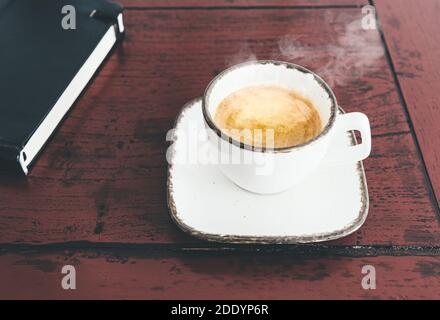 Nahaufnahme einer Tasse dampfenden heißen Espresso auf rustikalem Holz Schreibtisch mit Buch Stockfoto