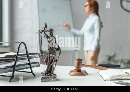 Themis Figur mit Gavel und Holzklotz auf Tisch mit Verschwommene Rechtsanwältin, die neben dem Flipchart auf dem Hintergrund steht Stockfoto