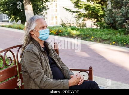 Älterer Mann in Schutzmaske sitzt auf einer Bank in Ein Stadtpark am sonnigen Herbsttag Stockfoto