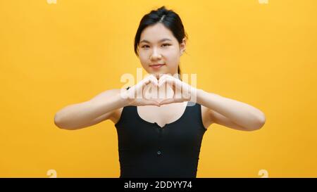 Schöne junge asiatische Frau macht Herz Form mit ihren Händen. Isoliert auf gelbem Hintergrund. Hochwertige Fotos Stockfoto
