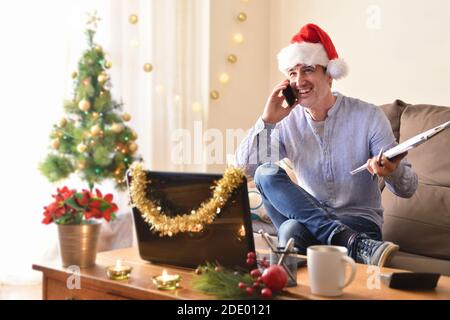 Lächelnder Mann, der am Telefon mit Weihnachtsmütze und Ordner in der Hand telefoniert und an den weihnachtsfeiertagen im Wohnzimmer zu Hause arbeitet. Horizontale Kompositio Stockfoto