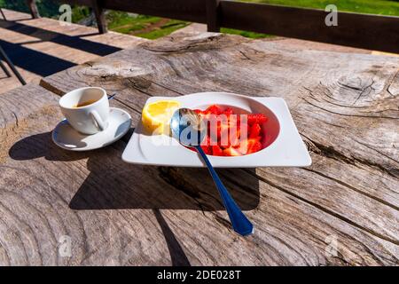 Weißer Tassenkaffee und kleiner runder Teller mit saftig leckeren Erdbeeren, eine Erdbeere liegt separat vor dem Fenster. Guten Morgen und gute Laune Stockfoto