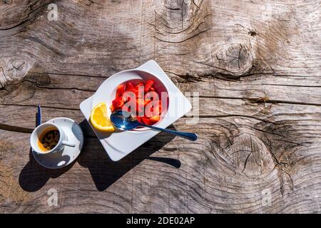 Weißer Tassenkaffee und kleiner runder Teller mit saftig leckeren Erdbeeren, eine Erdbeere liegt separat vor dem Fenster. Guten Morgen und gute Laune Stockfoto