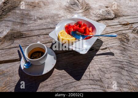 Weißer Tassenkaffee und kleiner runder Teller mit saftig leckeren Erdbeeren, eine Erdbeere liegt separat vor dem Fenster. Guten Morgen und gute Laune Stockfoto