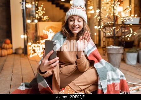 Junge Frau mit Hut und Karomutorum feiert allein Silvester Urlaub auf der Terrasse zu Hause, mit einem Videoanruf am Telefon. Konzept der Quarantäne und Selbstisolation während der Epidemie an Feiertagen Stockfoto