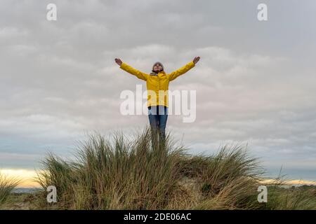 Frau trägt einen gelben Mantel auf einer Sanddüne mit erhobenen Armen, Erfolgskonzept. Stockfoto