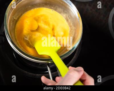 Kochen süßer Biskuitkuchen zu Hause - oben Ansicht der Rührmischung von Zutaten aus Honig, Zucker und rohen Eiern im Wasserbad mit Spatel auf dem Herd bei Stockfoto