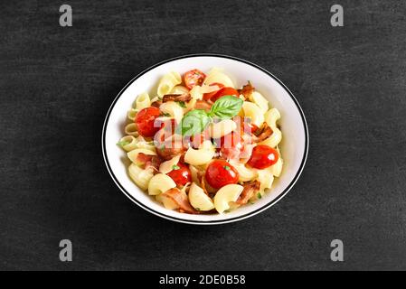 Pasta mit Speck, Tomaten, Parmesan-Käse und Basilikumblättern auf dem Teller auf schwarzem Stein Hintergrund mit freiem Textraum. Stockfoto