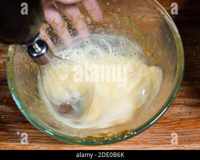 Kochen süßer Biskuitkuchen zu Hause - über Ansicht von Schlagen von rohem Ei in Glasschüssel durch elektrischen Mixer Auf alten Holztisch in der heimischen Küche (Mixer whi Stockfoto