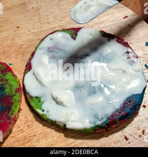 Kochen süßer Biskuitkuchen zu Hause - über Ansicht von Verbreitung von Sahne auf Schicht von bunten gebackenen Kuchen durch Spachtel auf alten Holzbrett in der heimischen Küche Stockfoto