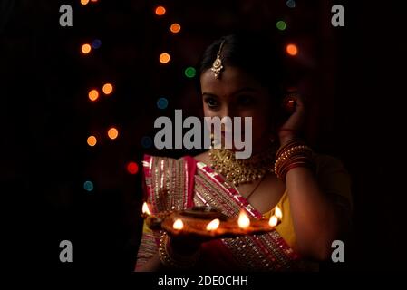 Eine junge und schöne indische Bengalin in indischer Tracht hält eine Diwali diya/Lampe in der Hand vor bunten Bokeh-Lichtern. Stockfoto