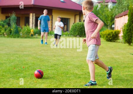 Zwei glückliche Söhne, die mit ihrem Vater im Garten Fußball spielen In der Nähe des Modern House Stockfoto