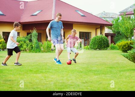 Zwei glückliche Söhne, die mit ihrem Vater im Garten Fußball spielen In der Nähe des Modern House Stockfoto