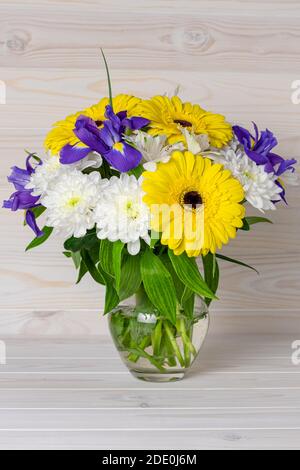 Ein Blumenstrauß in einem Glaskrug auf hellem Holzhintergrund. Stillleben im rustikalen Stil. Gelbe Gerbera, blaue Iris und weiße Chrysanthem Stockfoto