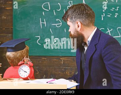 Junger männlicher Lehrer führt sein Kind Schüler zum Lernen, während Sohn schaut auf Kreidetafel mit Kritzeleien auf, sitzen im Klassenzimmer. Stockfoto