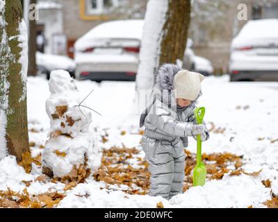 Glückliches Kind auf einem Winterspaziergang im Park. Ein lustiges Mädchen in einem silbernen Jumpsuit hält eine Spielzeugschaufel und modelliert einen Schneemann. Trockene Ahornblätter schauen heraus Stockfoto