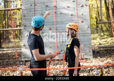Gut ausgestattet mit Sicherheitsausrüstung Mann und Frau stehen vor der Kletterwand im Vergnügungspark, bereit zum Klettern Stockfoto
