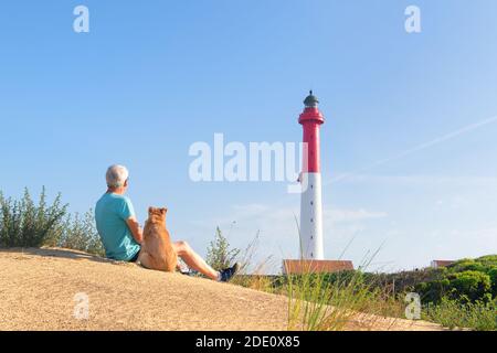 Mann und Hund sitzen auf Düne und schauen zu Leuchtturm rot und weiß am französischen Weststrand Stockfoto