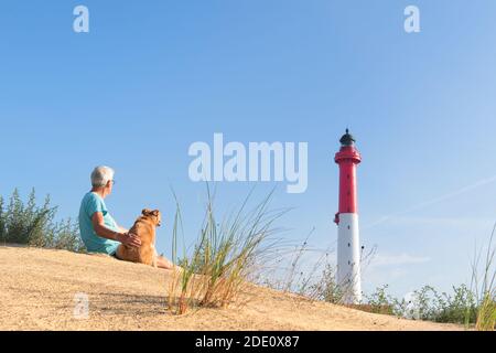 Mann und Hund sitzen auf Düne und schauen zu Leuchtturm rot und weiß am französischen Weststrand Stockfoto
