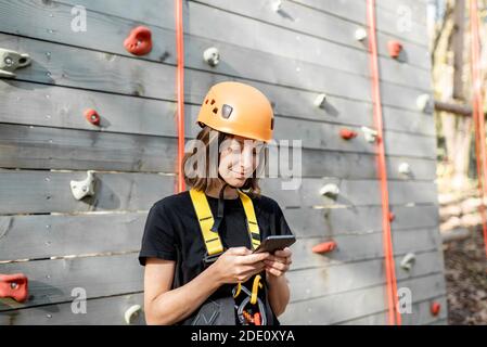 Junge Frau in schützender Sportausrüstung mit einem Smartphone In der Nähe der Kletterwand im Freien Stockfoto