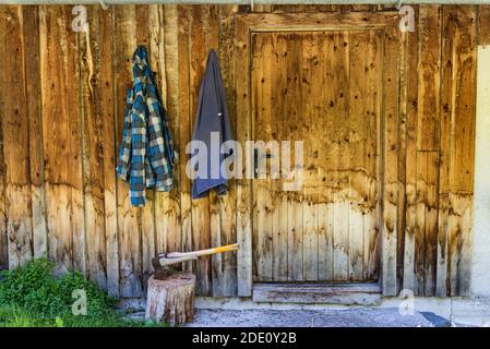 Zwei Holzachsen stecken im Holzhackkoffer. Im Hintergrund eine Holzwand des Hauses und zwei Arbeitskleidung, die an einem Haken hängen. Stockfoto