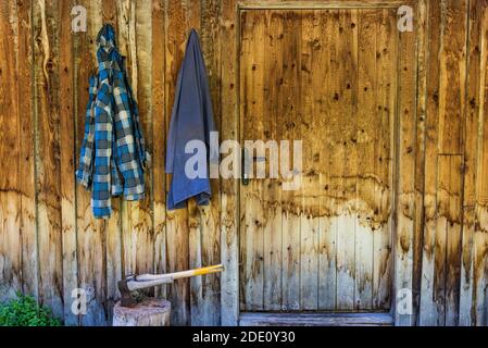 Zwei Holzachsen stecken im Holzhackkoffer. Im Hintergrund eine Holzwand des Hauses und zwei Arbeitskleidung, die an einem Haken hängen. Stockfoto