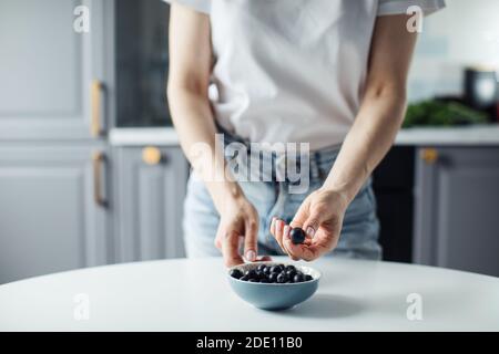 Die Hände der Frau nehmen Blaubeeren vom Teller. Auf dem Hintergrund einer schönen Küche. Stockfoto