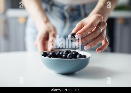 Die Hände der Frau nehmen Blaubeeren vom Teller. Auf dem Hintergrund einer schönen Küche. Stockfoto