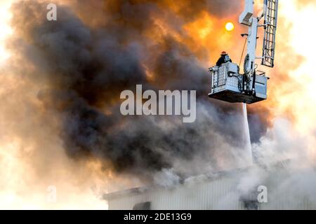 Feuerwehrmann, der einen Gebäudebrand vom Eimer über dem Gebäude bekämpft. Ein Schlauch sprüht Wasser durch den Rauch. Feuerwehrmann trägt Atemgerät. Stockfoto