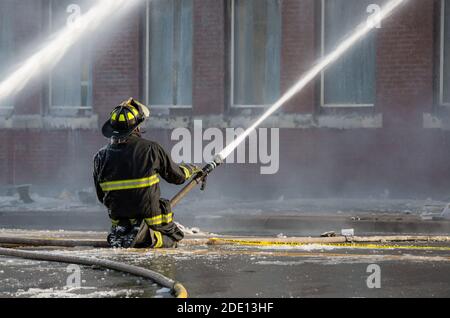 Ein Feuerwehrmann kniet mitten auf nasser, eisiger Straße. Er verwendet einen Schlauch, um Wasser zu sprühen. Die Löschschläuche befinden sich am Boden. Das Gebäude ist im Hintergrund. Stockfoto