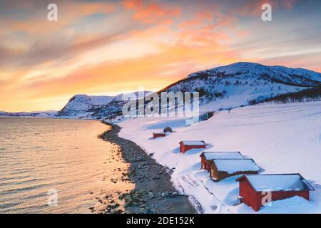 Sonnenuntergang über roten Hütten im Schnee entlang Porsangerfjord mit Nordkap (Nordkapp) im Hintergrund, Troms Og Finnmark, Arktis, Norwegen Stockfoto