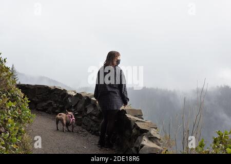 Eine junge Frau in einer Gesichtsmaske und ihr Hund, der die Aussicht vom Cape Perpetua Aussichtspunkt in Oregon, USA, betrachtet. Stockfoto