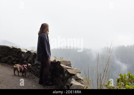 Eine junge Frau in einer Gesichtsmaske und ihr Hund, der die Aussicht vom Cape Perpetua Aussichtspunkt in Oregon, USA, betrachtet. Stockfoto