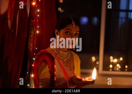 Eine junge und schöne indische Bengalin in indischer Tracht hält eine Diwali diya/Lampe in der Hand vor bunten Bokeh-Lichtern. Stockfoto