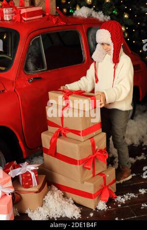 Junge auf dem Hintergrund eines roten Autos mit Geschenken. Kind baut einen Turm aus Weihnachtskisten. Stockfoto