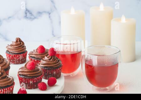 Schokolade Cupcakes mit Schokolade Buttercreme Zuckerguss mit frischen Himbeeren verziert, und Himbeer-Tee in Glas Tassen schließen auf hellem Hintergrund. Stockfoto