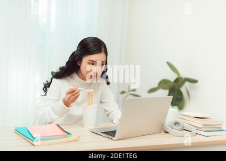 Asien freiberufliche Geschäftsfrau essen Instant Nudeln während der Arbeit an Laptop im Wohnzimmer im Heimbüro Stockfoto