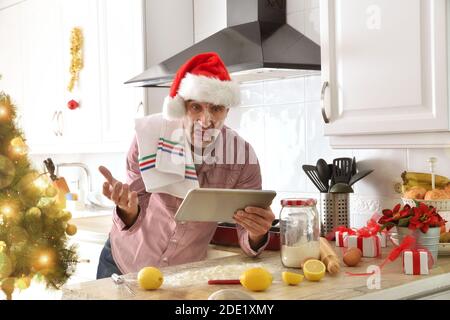 Ein verärgerter Anfänger Koch Blick auf eine digitale Tablette Rezept Mit weihnachtshut mit Zutaten auf der Küchenbank und Baumhintergrund mit Lichtern Stockfoto