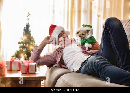 Mann liegt zu Hause auf dem Sofa mit seinem Hund in Weihnachtskostüm mit geschmückten Baum und Geschenken gekleidet. Stockfoto