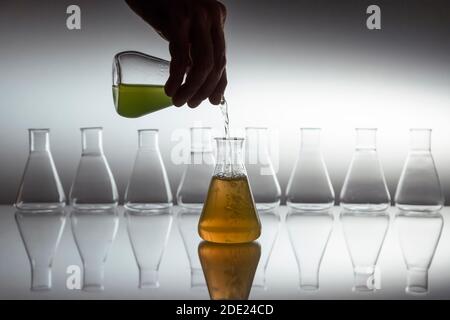 Hand Gießen grün in orange Flüssigkeit in wissenschaftlichen Labor Glas erlenmeyerkolben mit Glasgeräten auf reflektierende Oberfläche. Stockfoto